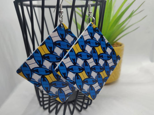 Pair of blue and yellow patterned earrings on a black stand with a plant in the background.